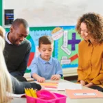 In this image, a teacher engages with a young student and their parents during a parent-teacher conference in a welcoming classroom environment. The teacher shares insights, likely discussing the student's progress while using learning materials as visual aids. The colorful classroom decor, including educational posters and charts, enhances the setting and creates a positive atmosphere. This moment captures the essence of collaboration between educators and families, focusing on student success and fostering a strong partnership for academic growth.