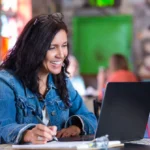 Smiling teacher writing notes while working on a laptop.