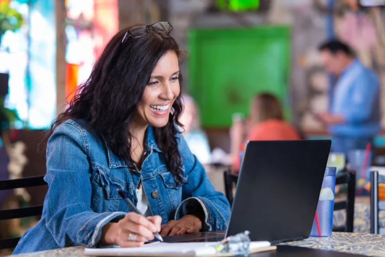 Smiling teacher writing notes while working on a laptop.