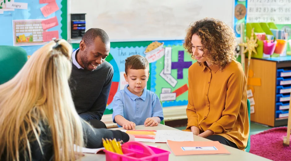 In this image, a teacher engages with a young student and their parents during a parent-teacher conference in a welcoming classroom environment. The teacher shares insights, likely discussing the student's progress while using learning materials as visual aids. The colorful classroom decor, including educational posters and charts, enhances the setting and creates a positive atmosphere. This moment captures the essence of collaboration between educators and families, focusing on student success and fostering a strong partnership for academic growth.