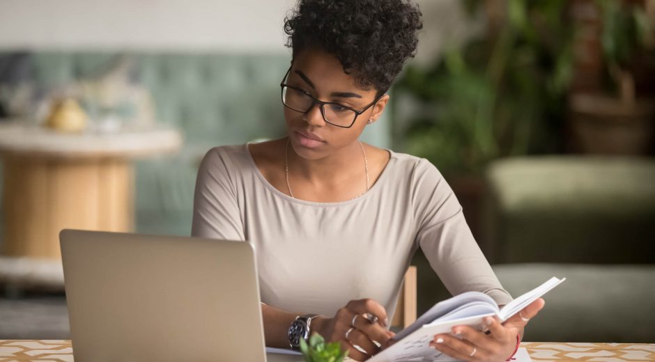Focused young african american businesswoman or student looking at laptop holding book learning, serious black woman working or studying with computer doing research or preparing for exam online