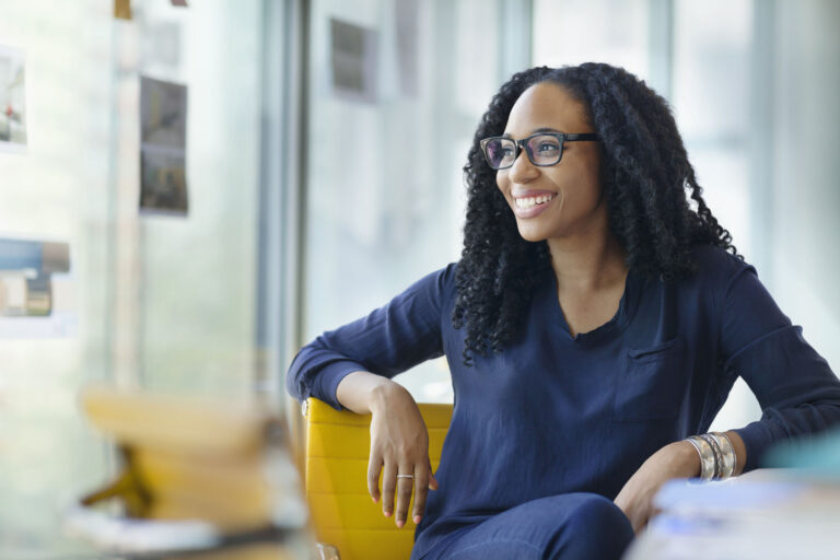Portrait of young Black female woman designer owner entrepreneur small business new hire intern smiling proud dedicated passionate looking at camera sitting at desk in creative design studio office startup company contemporary modern daylight bright sunny natural light curly hair eyeglasses