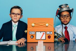 Two young boys and businessmen dressed in suits and glasses sit at a desk with a homemade lie detector machine. This serious interview is conducted in an office and the machine is predicting a lie from the job candidate. Dishonesty will eventually catch up to you.